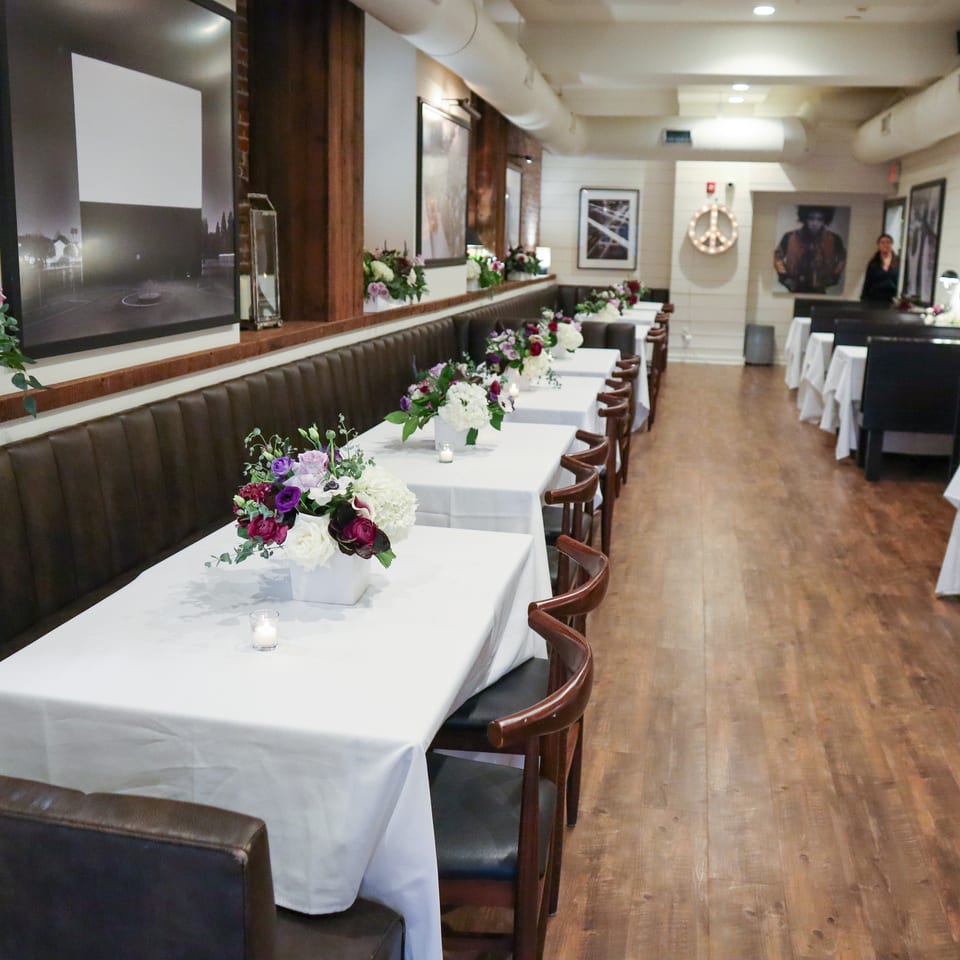 Elegant restaurant dining room with white tablecloths, floral centerpieces, dark leather banquettes, artwork, and warm wood floors.