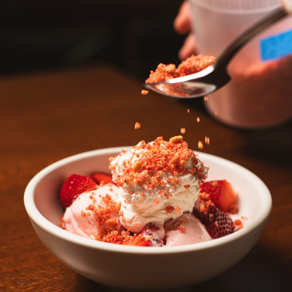 Bowl of ice cream topped with whipped cream, strawberries, and red crumble, while a spoon sprinkles additional topping.