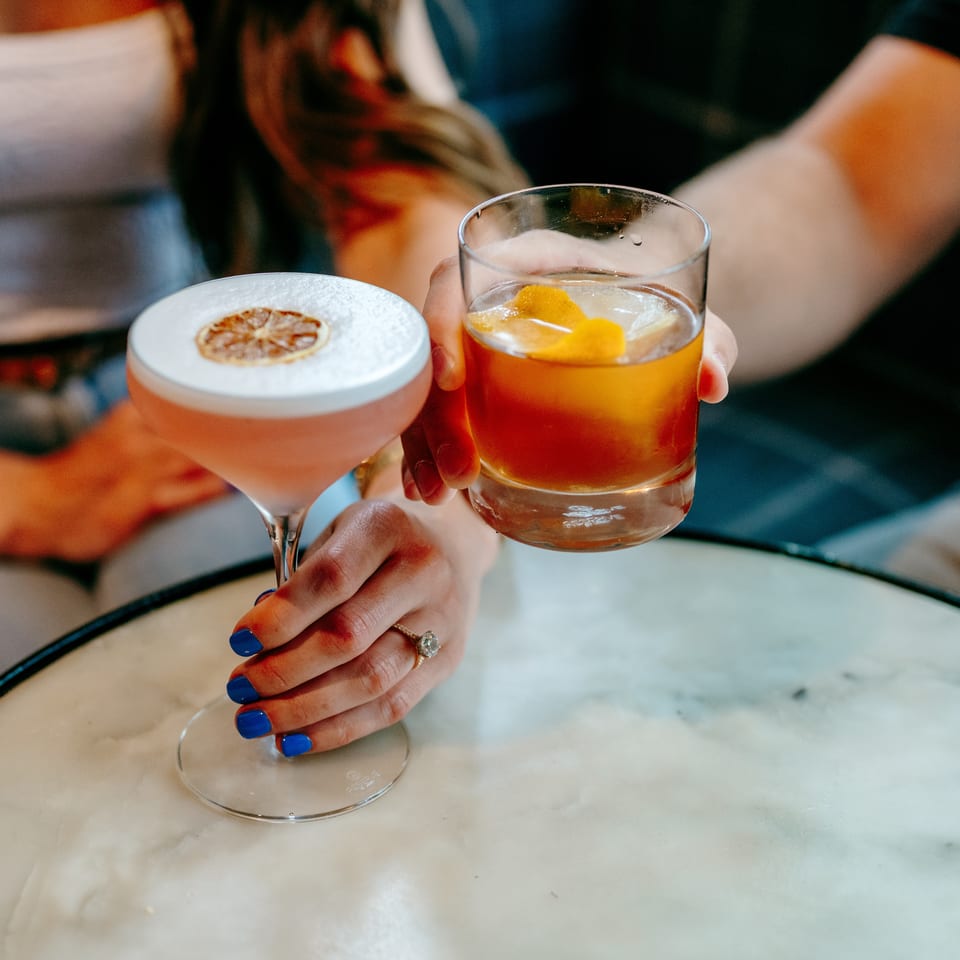 Two cocktails, one topped with foam and dried citrus, the other garnished with an orange peel, are held over a marble table.