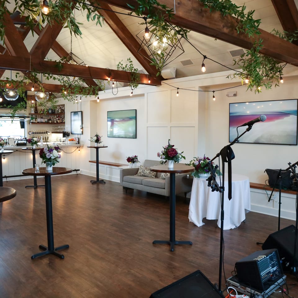 Restaurant event space with floral centerpieces, bar, greenery-covered beams, lounge seating, and microphones set up for live music.