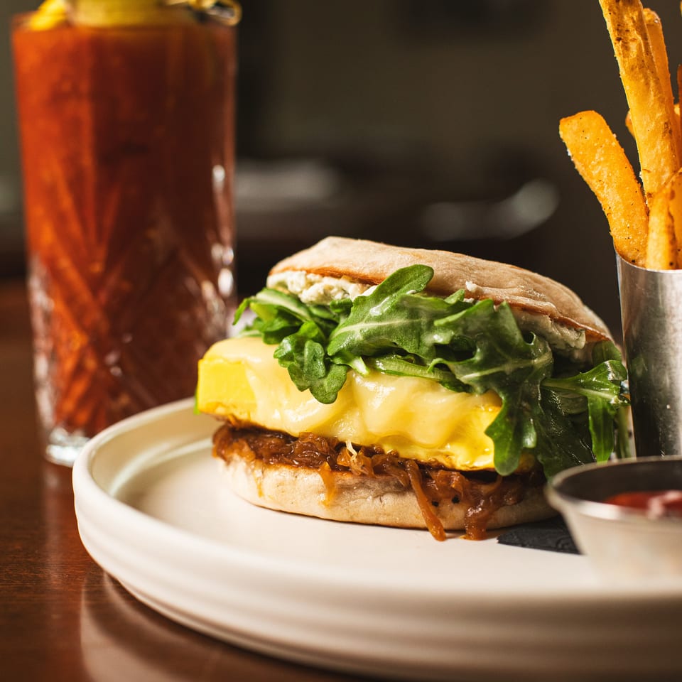 Cheeseburger topped with melted cheese, greens, and onions on a plate, accompanied by fries and a tomato-garnished drink.