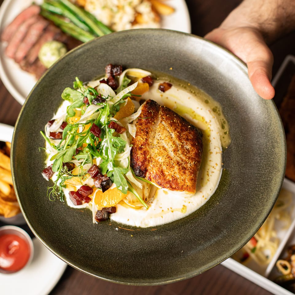 Seared fish with creamy sauce, citrus, greens, and diced vegetables on a dark plate, held above a restaurant table.