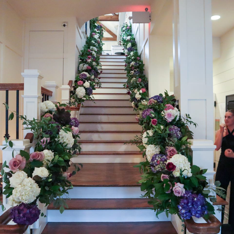 Grand wooden staircase adorned with abundant pink, white, and purple floral arrangements, with two women standing beside it.