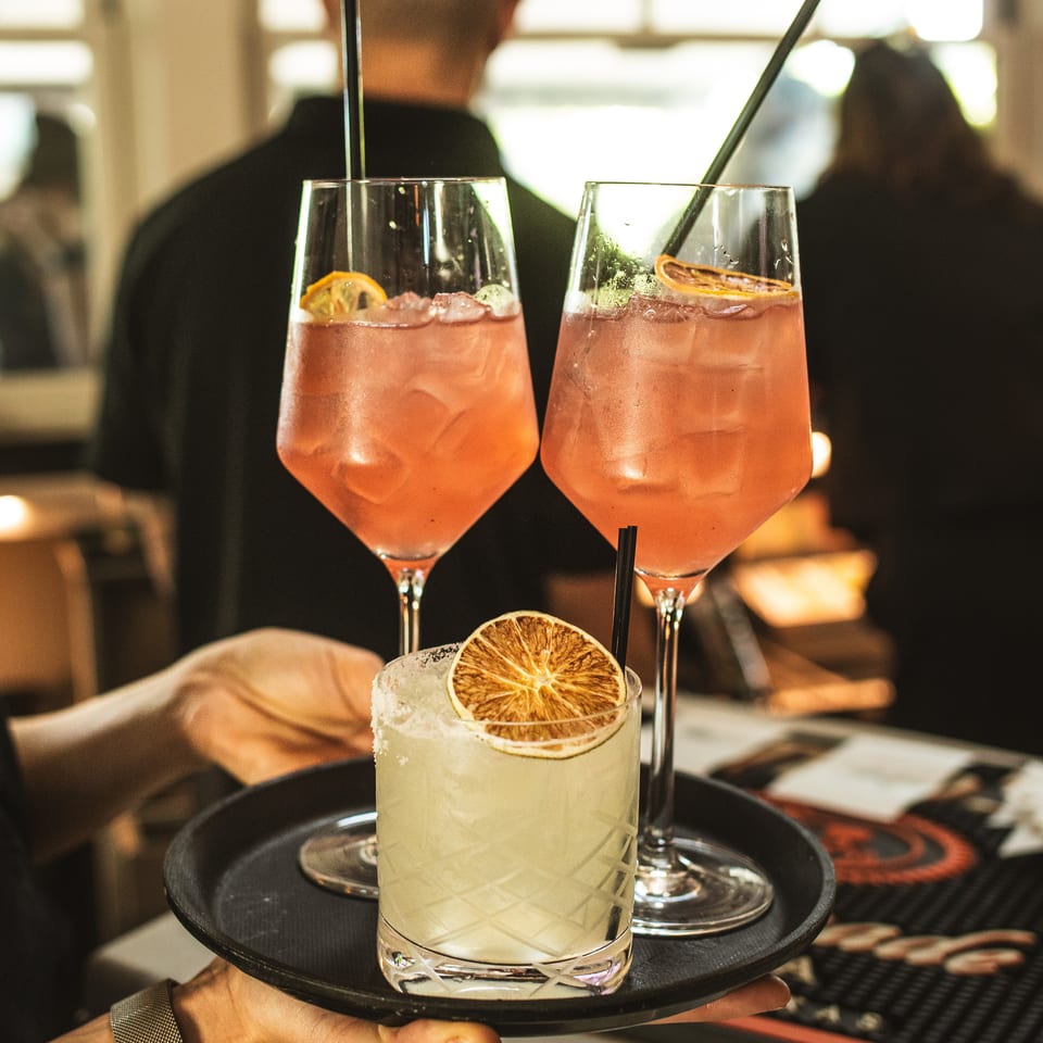 Server holds a black tray with three citrus-garnished cocktails in a warmly lit restaurant.