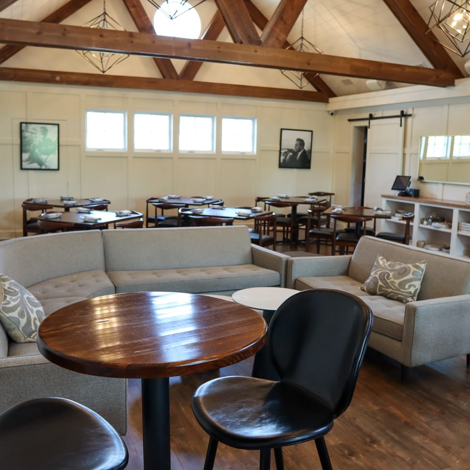 Bright restaurant dining room with exposed wooden beams, leather chairs, round tables, upholstered sofas, and framed artwork.