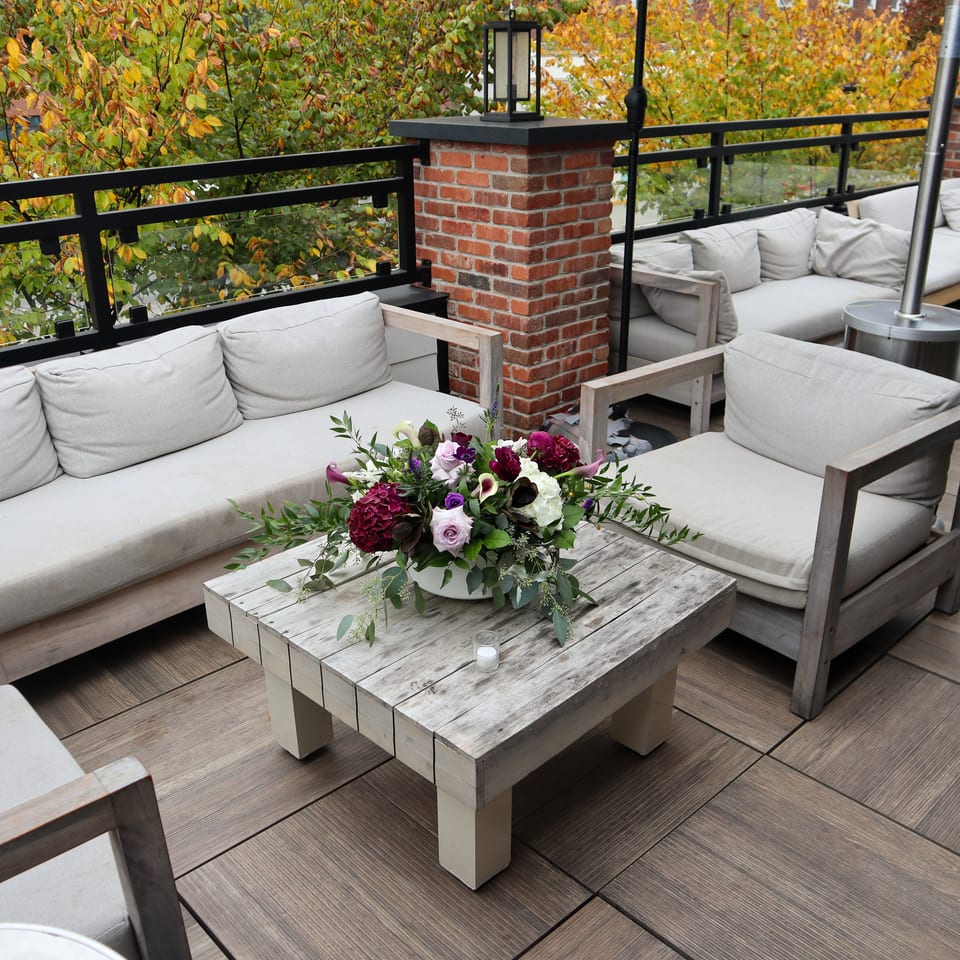 Outdoor restaurant patio with cushioned seating, a floral centerpiece, brick columns, lanterns, and autumn foliage.