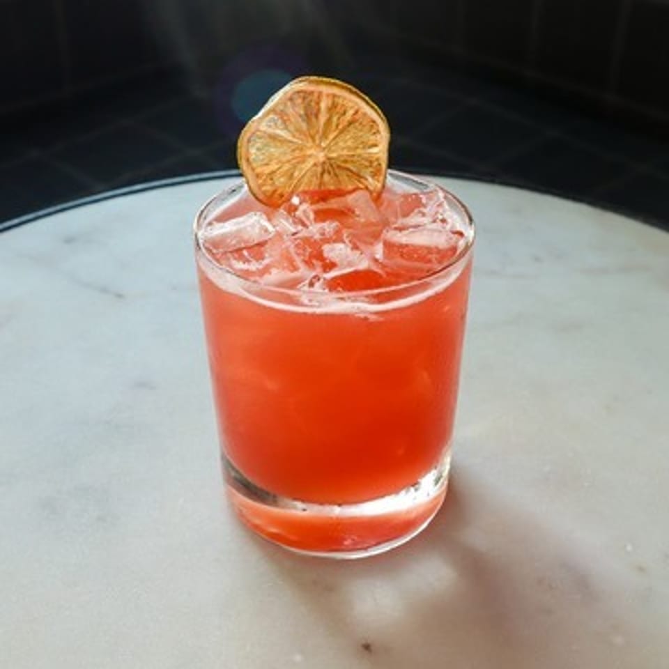 Bright red cocktail with ice in a short glass, garnished with a dried citrus wheel on a marble tabletop.