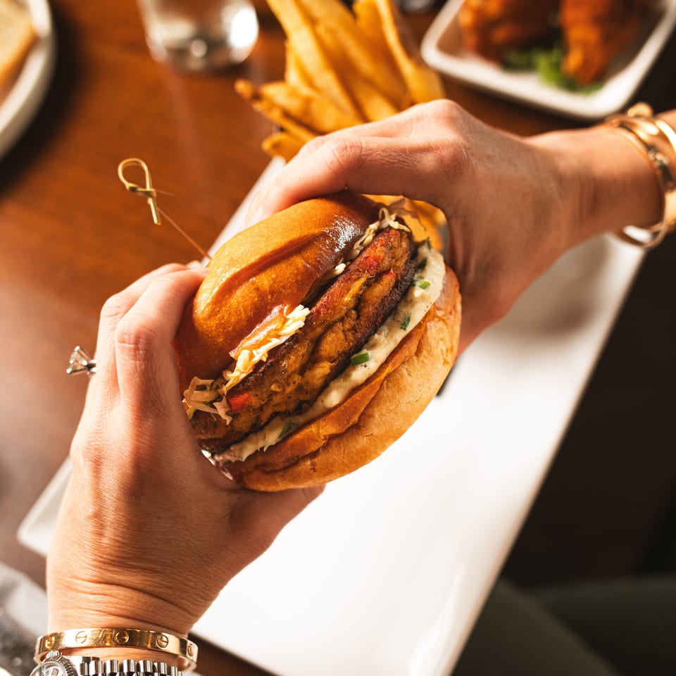 Hand holding a loaded burger with crispy fried filling and sauce, accompanied by golden fries on a restaurant table.