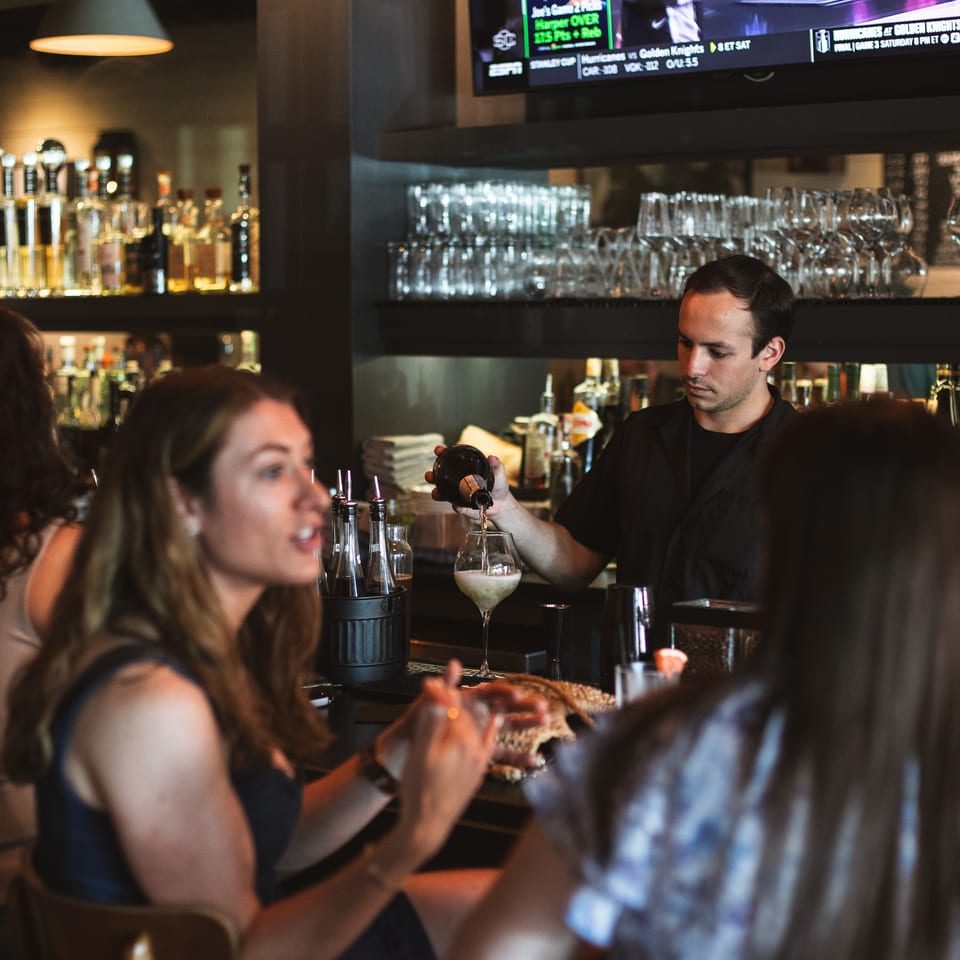 Bartender pours a drink at a lively restaurant bar, surrounded by glassware, bottles, diners, and a sports television.