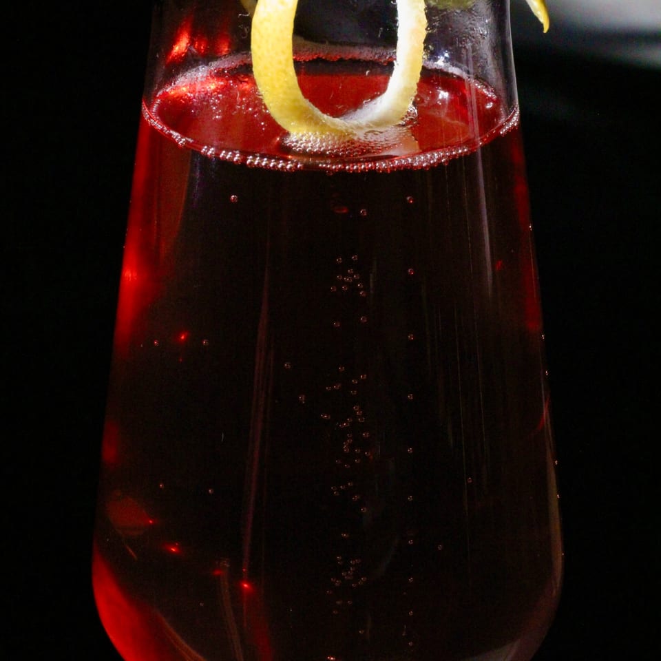 Dark red cocktail in a stemmed glass, garnished with a long curled citrus peel.