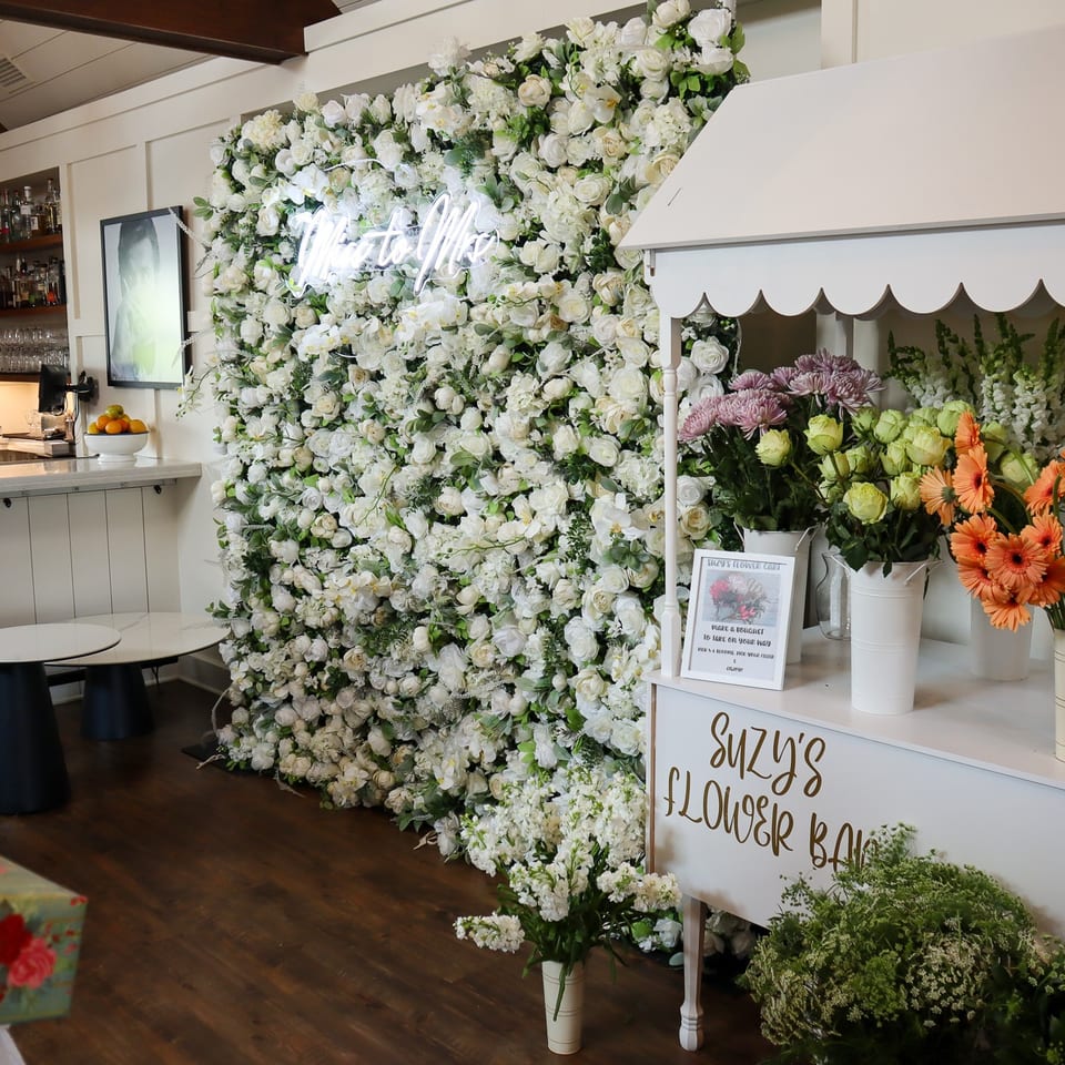 Restaurant interior featuring a flower market display, white floral wall, colorful bouquets, bar shelves, and a neon sign.