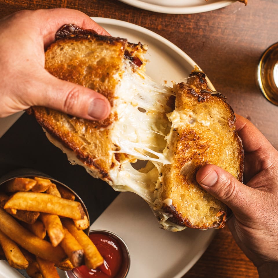Hands pull apart a grilled, cheese-filled sandwich over fries with ketchup on a restaurant table.